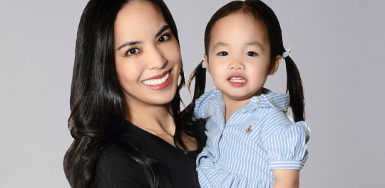 A woman with long black hair is holding a young child with pigtails wearing a striped shirt. Both are smiling against a plain background.