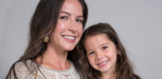 A woman and a young girl smiling at the camera against a plain background.