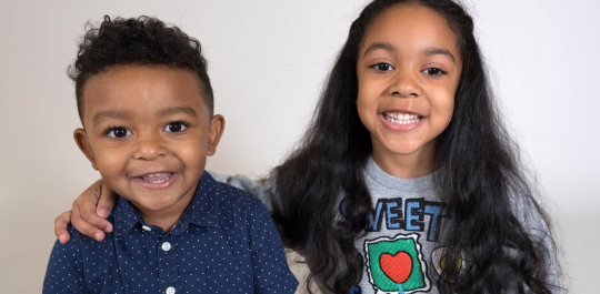 Two smiling children pose together; the young boy wears a blue polka dot shirt, and the girl with long hair wears a gray sweater with a colorful design.