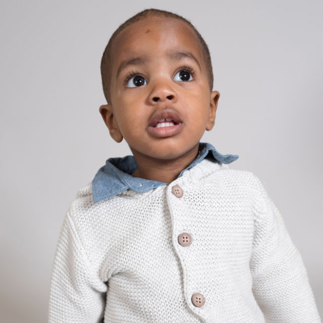 A toddler wearing a light-colored knit sweater with a blue collar looks upward against a plain background.