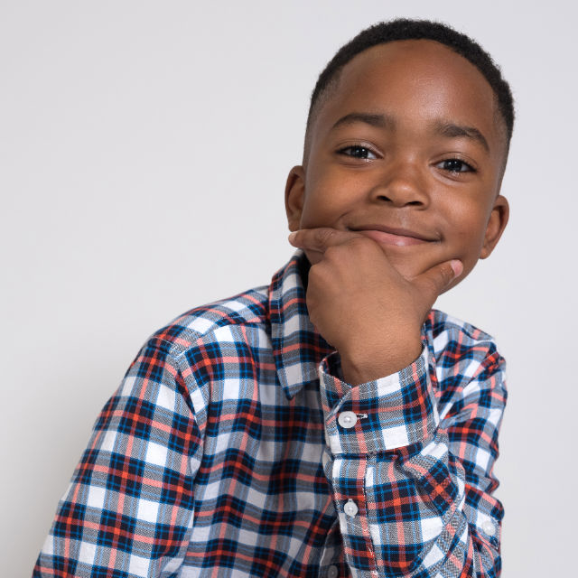 Child in a plaid shirt smiling with hand on chin against a plain background.