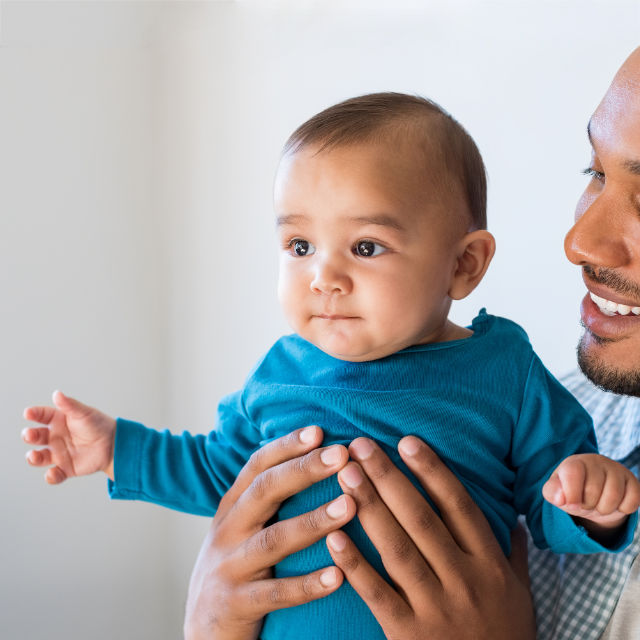Adult holding a baby wearing a blue shirt. The adult is smiling and looking at the child. The baby is looking to the side.