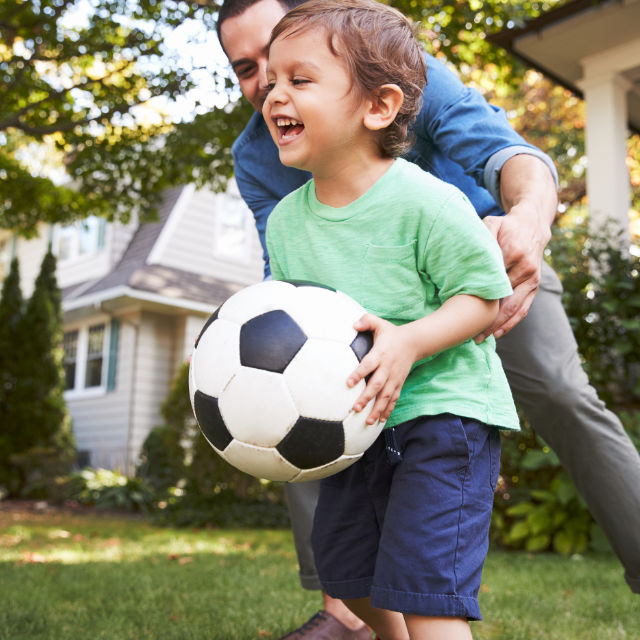 Child holding a soccer ball, smiling with an adult crouching behind, in a grassy backyard with a house and trees in the background.