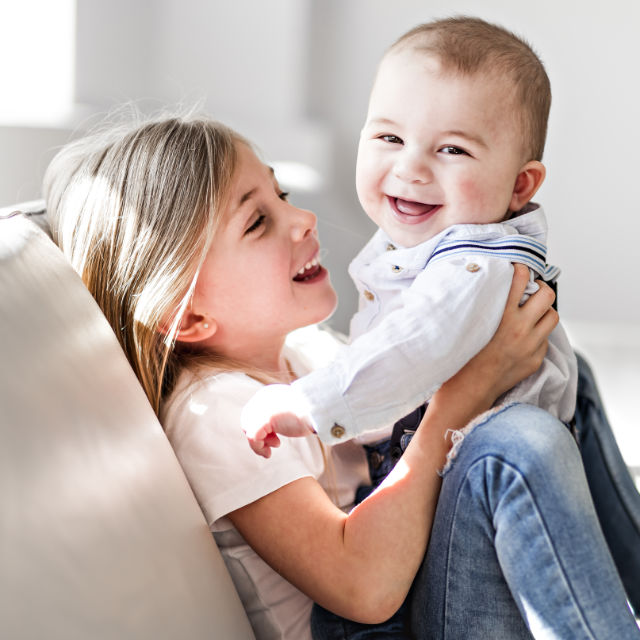 A young girl with long hair happily holds a laughing baby wearing a white shirt. Both are sitting on a couch in a brightly lit room.