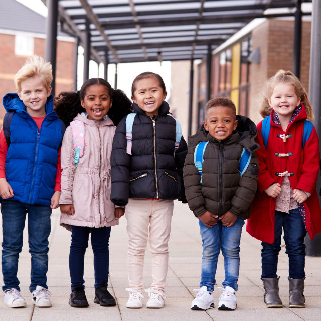 Five children smiling, wearing jackets and backpacks, stand in a row outdoors under a covered walkway.