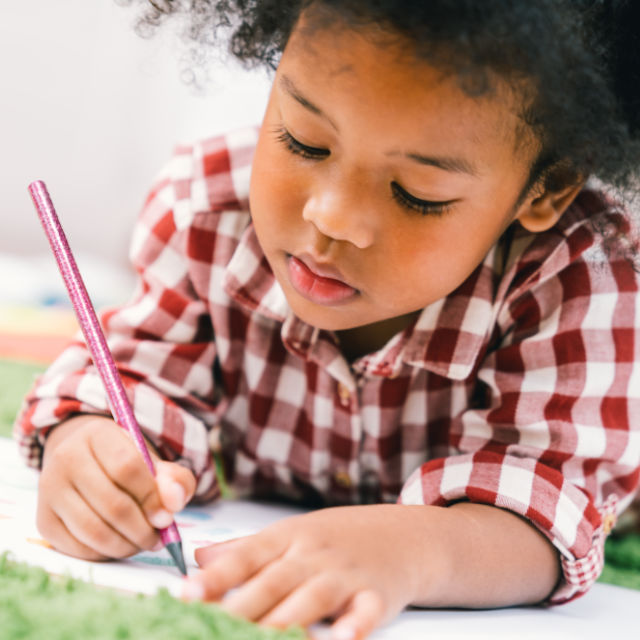 Child in a plaid shirt is drawing with a pencil on paper, focused intently on their work.
