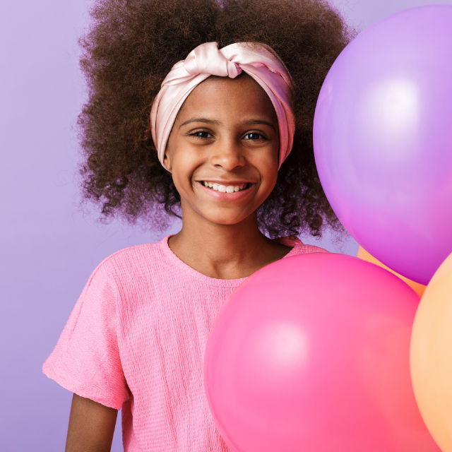 Smiling child in a pink top and headband holds colorful balloons against a purple background.