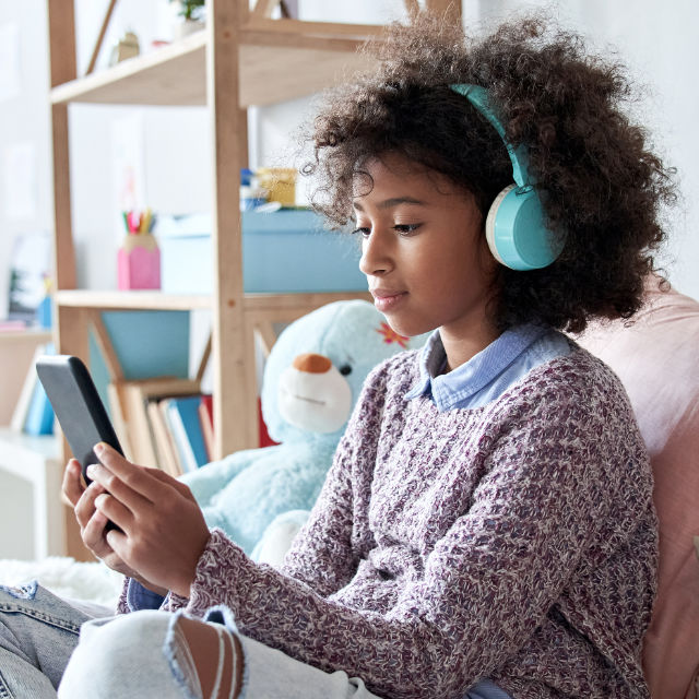 A young girl wearing headphones looks at a smartphone while sitting on a couch in a room with a teddy bear and shelves in the background.