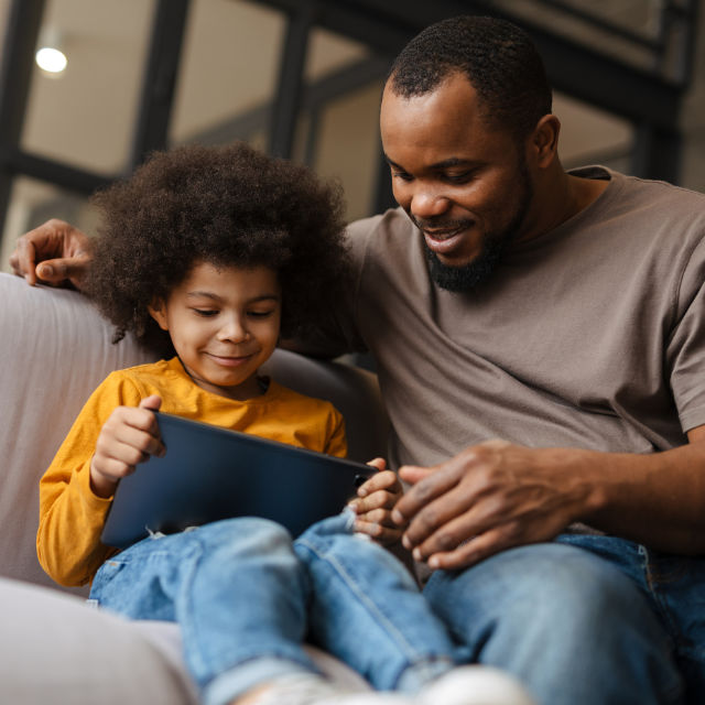 A man and a child sit on a couch, smiling while looking at a tablet.