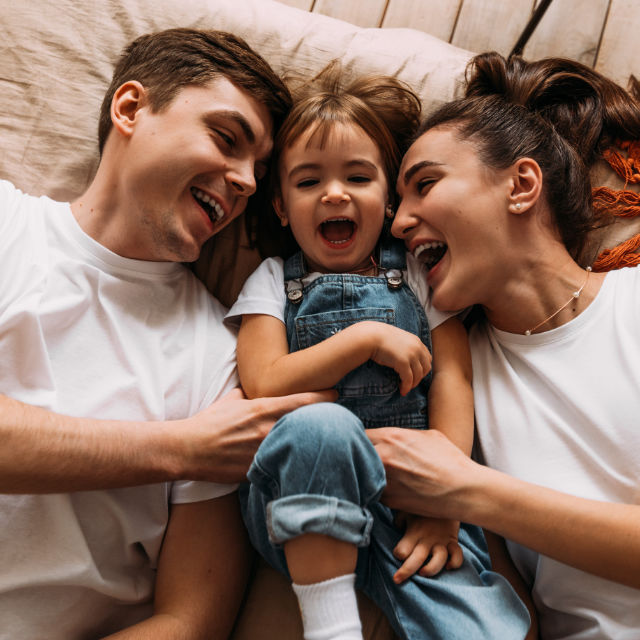 A family of three lies on a bed, smiling and laughing. The young child in the middle wears denim overalls. The adults on either side wear white shirts.