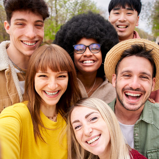 Group selfie of six smiling people outdoors, featuring diverse clothing styles and backgrounds.
