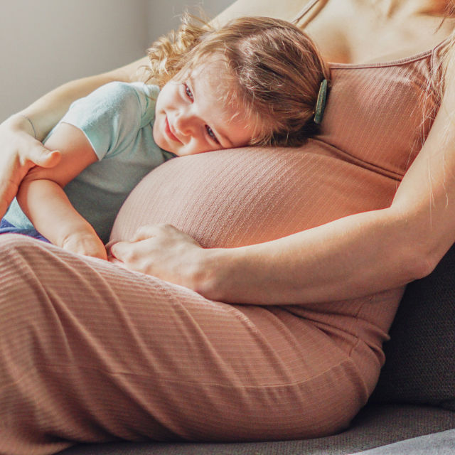 A young child rests on the belly of a pregnant person sitting on a couch, wearing a pink dress.