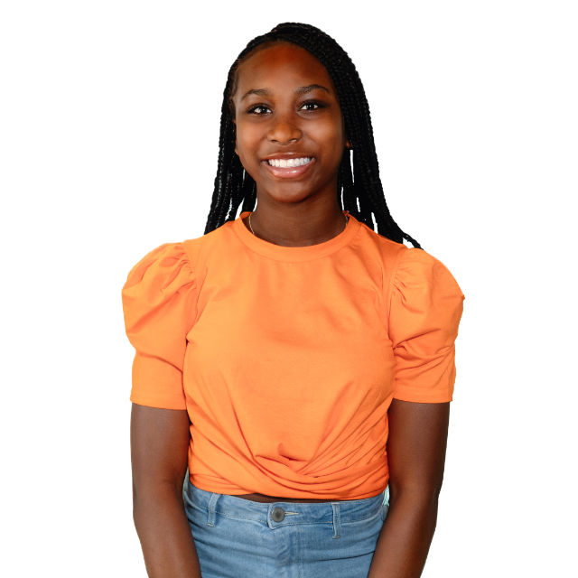 Smiling person with braided hair wearing an orange top and jeans, standing against a white background.