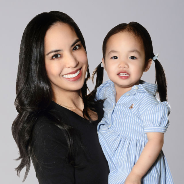 A woman with long dark hair holds a young girl with pigtails, both smiling, against a neutral background. The girl is wearing a blue striped dress.