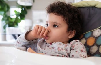 Young child with curly hair and a floral shirt sits in a high chair, sucking their thumb and looking to the side in a bright, indoor setting.