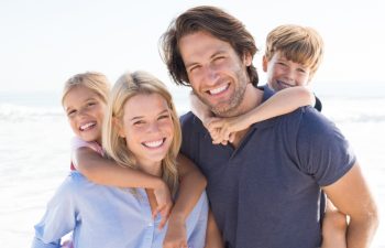 Parents giving piggyback ride to kids at beach. Close up of smiling family having fun at summer vacation. Portrait of happy family looking at camera at beach.