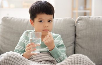 An Asian little boy sitting on a couch with a glass of water in one hand and a pill in the other hand